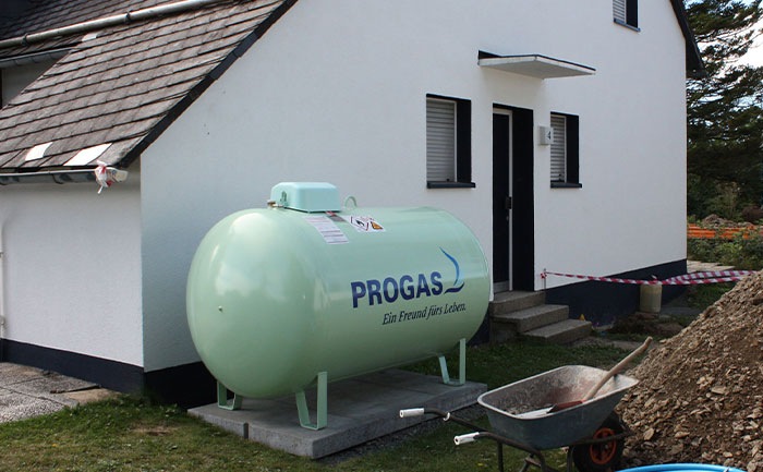 Ein PROGAS-Flüssiggastank steht vor einem Einfamilienhaus. Neben dem Haus und dem Tank sieht man eine kleine Baustelle mit Absperrband, Schubkarre und aufgeschütteter Erde.