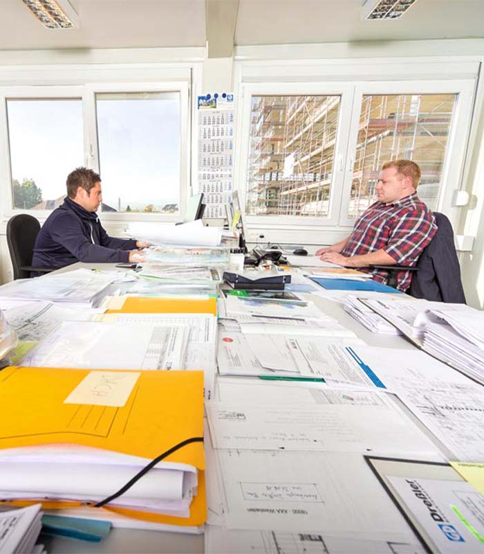 Das Foto zeigt Bauleiter Sebastian Hessler im Gespräch mit einem anderen Mann. Sie sitzen an einem Tisch in einem Baucontainer. 