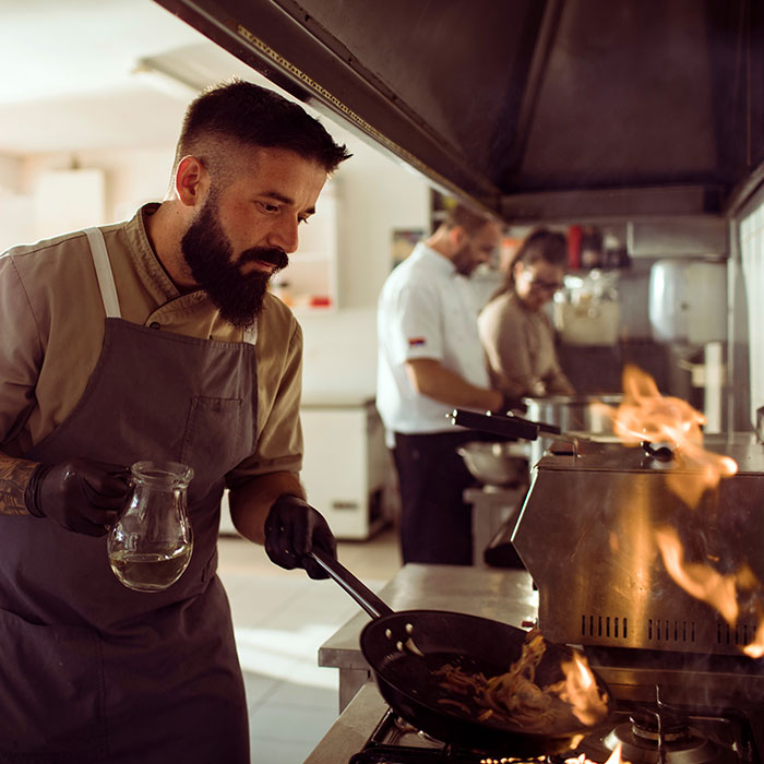 Foto eines Profikochs in der Gastronomie, der auf einem Flüssiggas Gasherd auf einer Flamme eine Pfanne schwenkt.