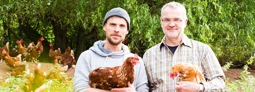 Foto zweier Hühnerbauern vor einem Wäldchen. In ihrer Hand halten die beiden je ein glückliches Huhn, im Hintergrund laufen noch mehr herum.