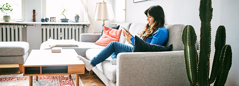 Eine junge Frau in einem modernen Wohnzimmer. Sie liest etwas am Handy und hat es sich auf dem Sofa bequem gemacht. Im Hintergrund ist eine warme Heizung.