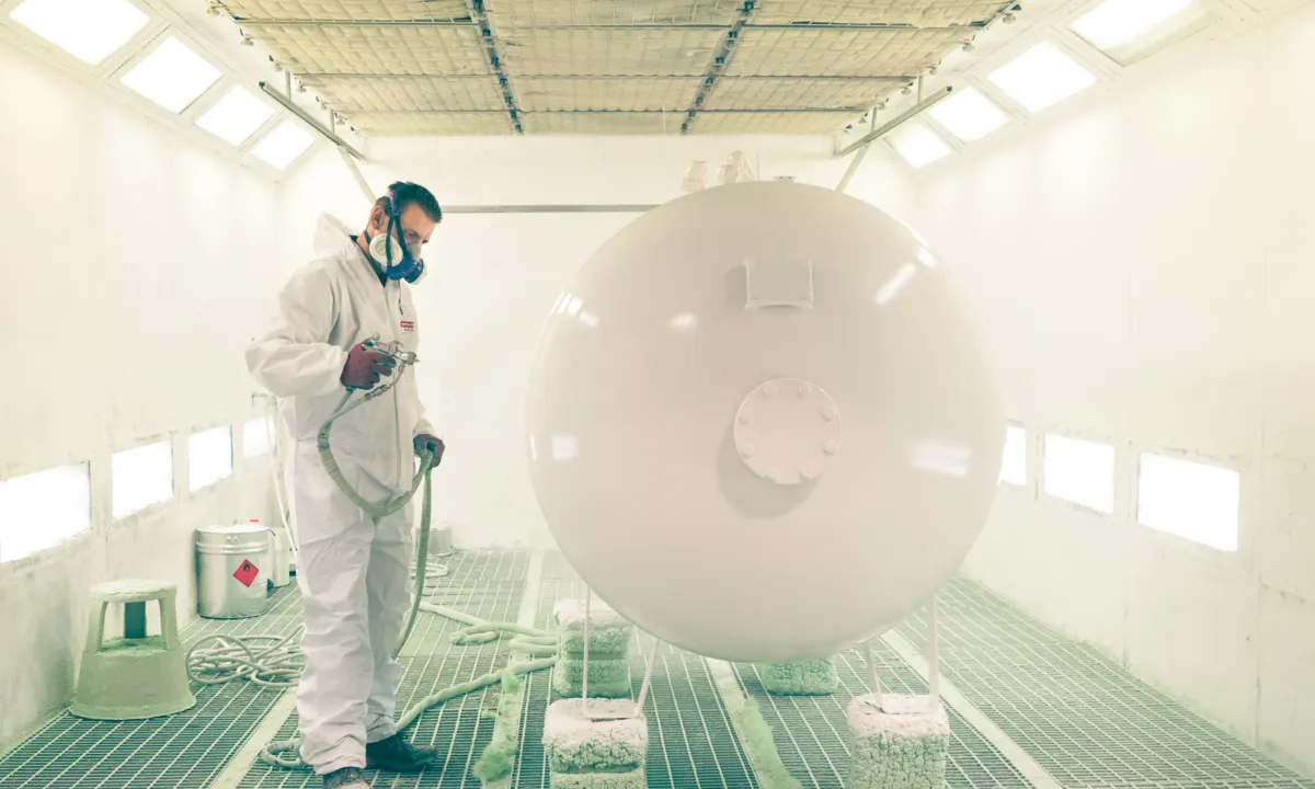 Ein Mann in Schutzkleidung lackiert einen Flüssiggastank, der in der Fabrik steht.