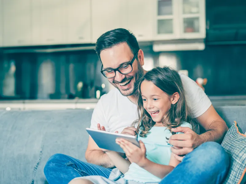 Ein junger Vater und seine kleine Tochter sitzen mit dem Tablet auf der Couch und freuen sich über die Angebote in der PROGAS-Vorteilswelt.